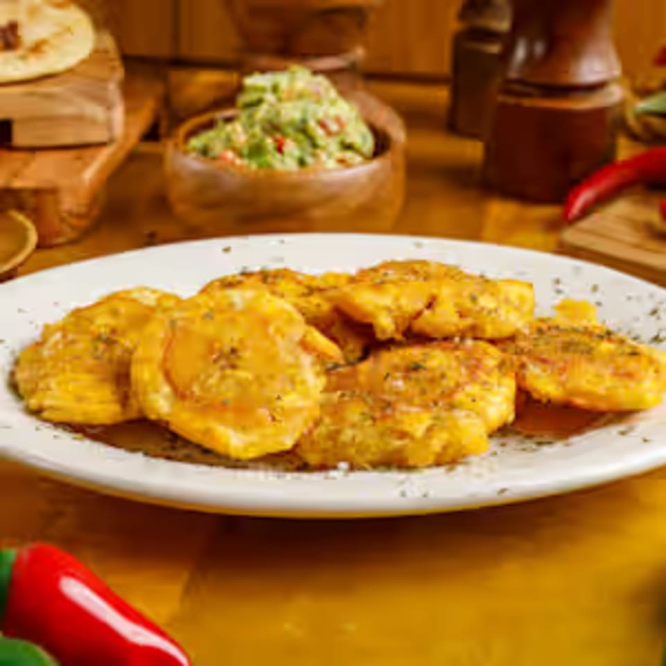 A plate of golden fried plantains, seasoned with herbs, with guacamole and tortillas in the background.