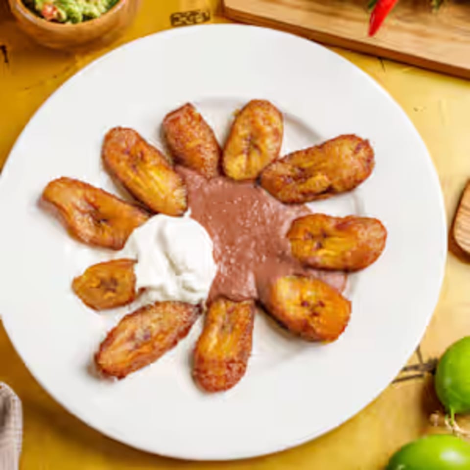 Delicia's fried plantains arranged in a circle around refried beans and sour cream on a white plate.