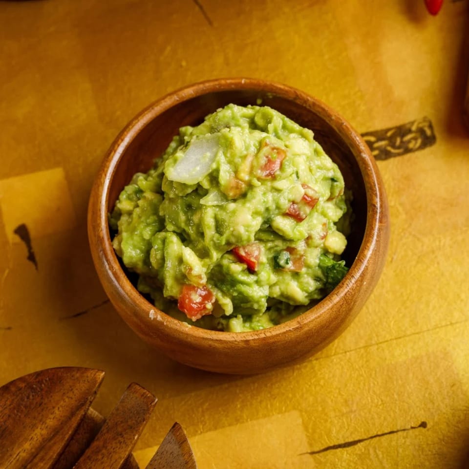 A wooden bowl filled with chunky green guacamole, diced tomatoes, and onions on a golden surface.