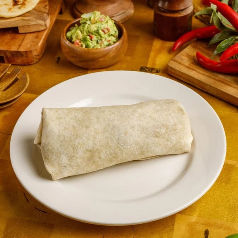 A large burrito on a white plate, with guacamole and red chilies in the background.